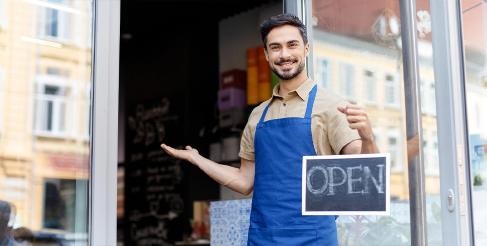Smiling small business owner in apron holding an ‘Open’ sign at the entrance of a shop